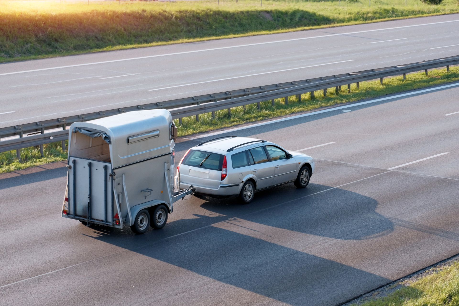 Silver car towing a horse trailer on a highway, with road barriers and grassy fields nearby.