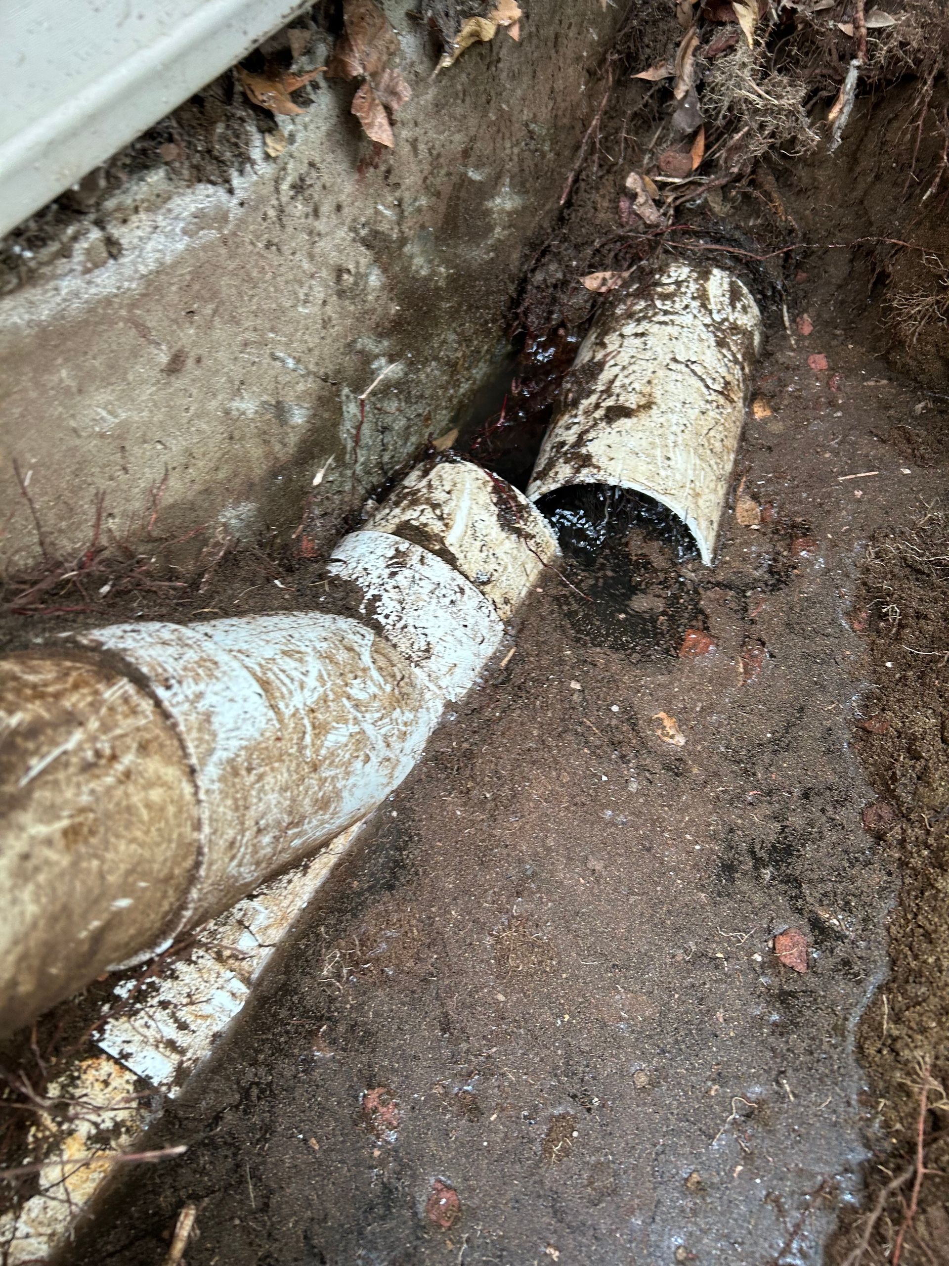 Broken white pipe leaking into muddy ground near a foundation wall