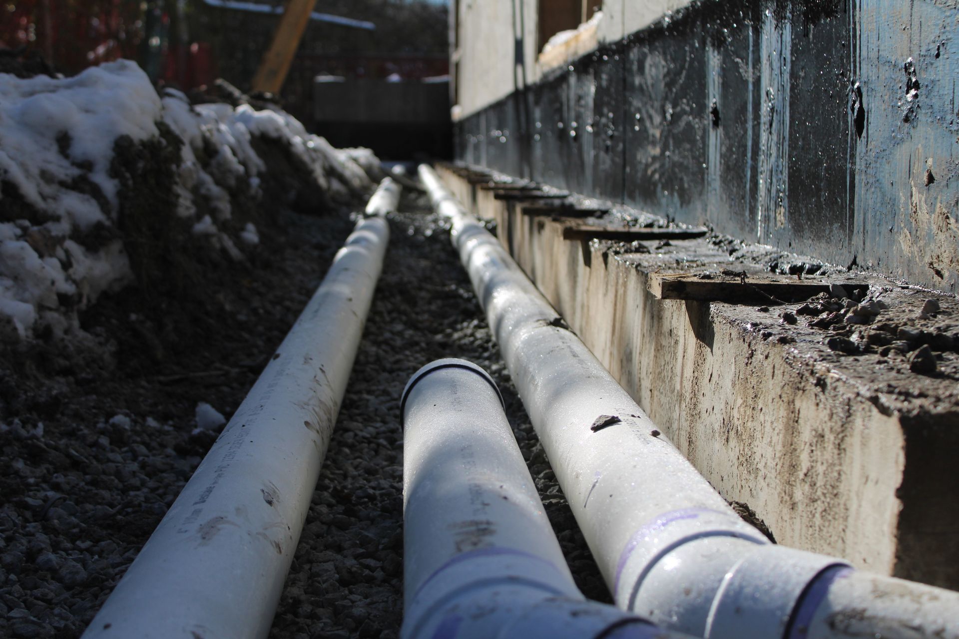 Snow-covered newly installed PVC pipes, running beside a concrete foundation of a newly constructed home