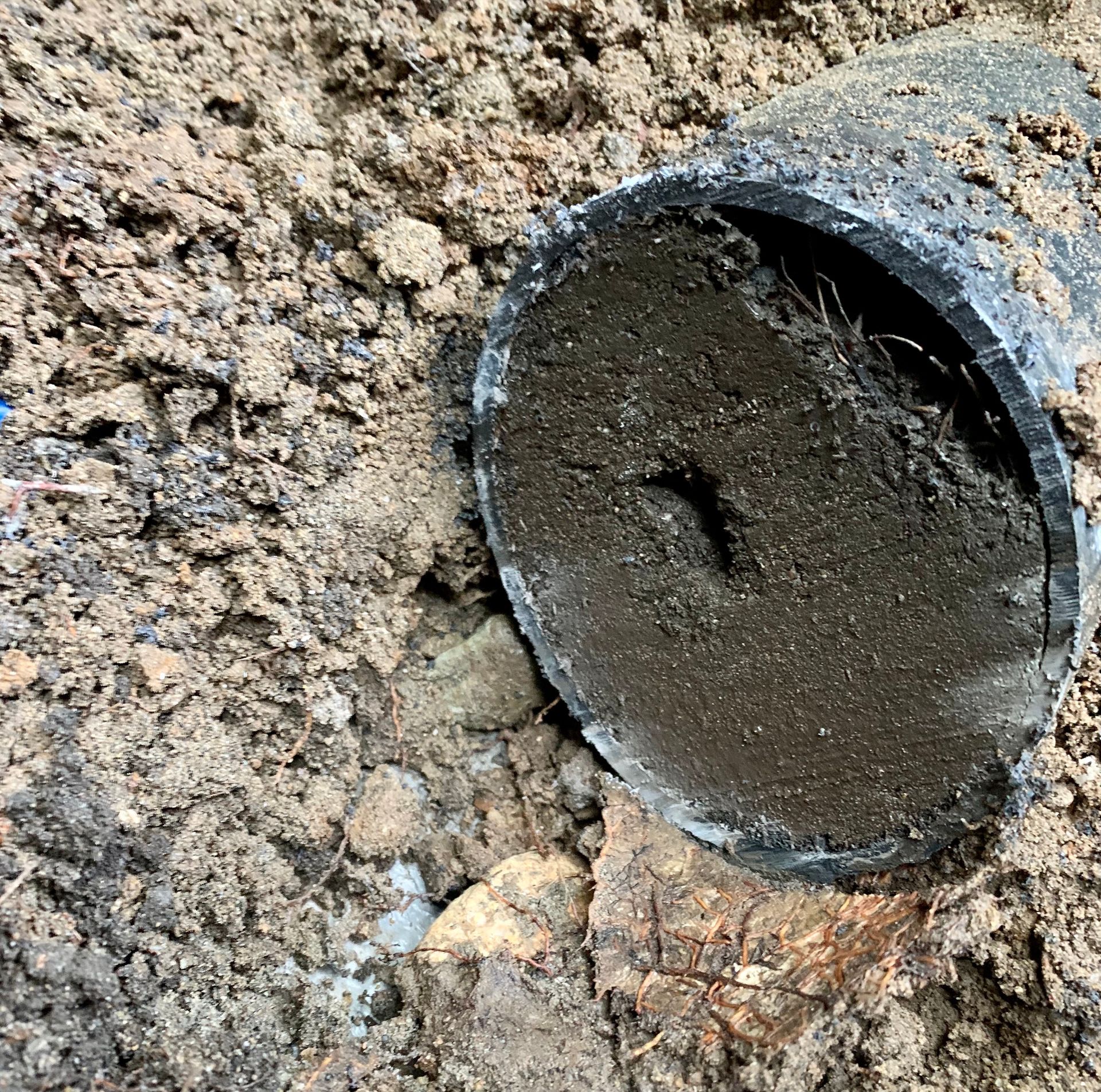 A black pipe opening clogged with mud, set in rocky, heavy clay soil.