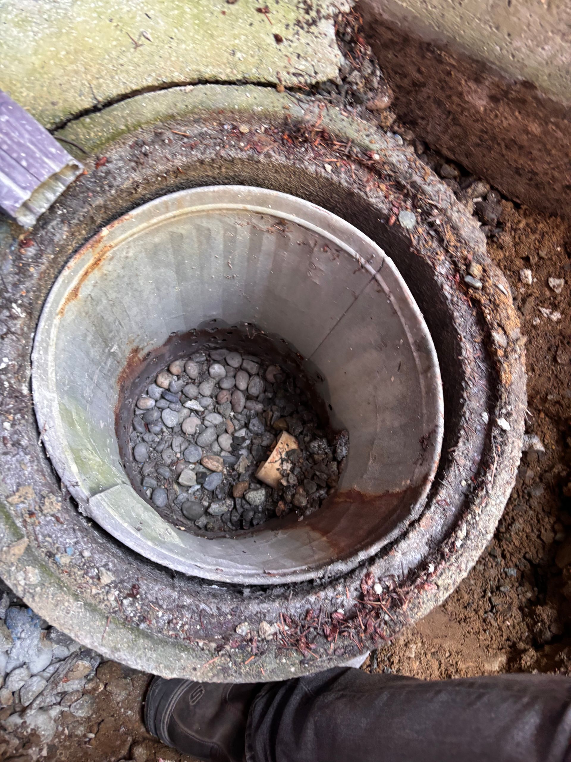 Open original sump  with a metal can insert and gravel inside, surrounded by dirt and concrete.