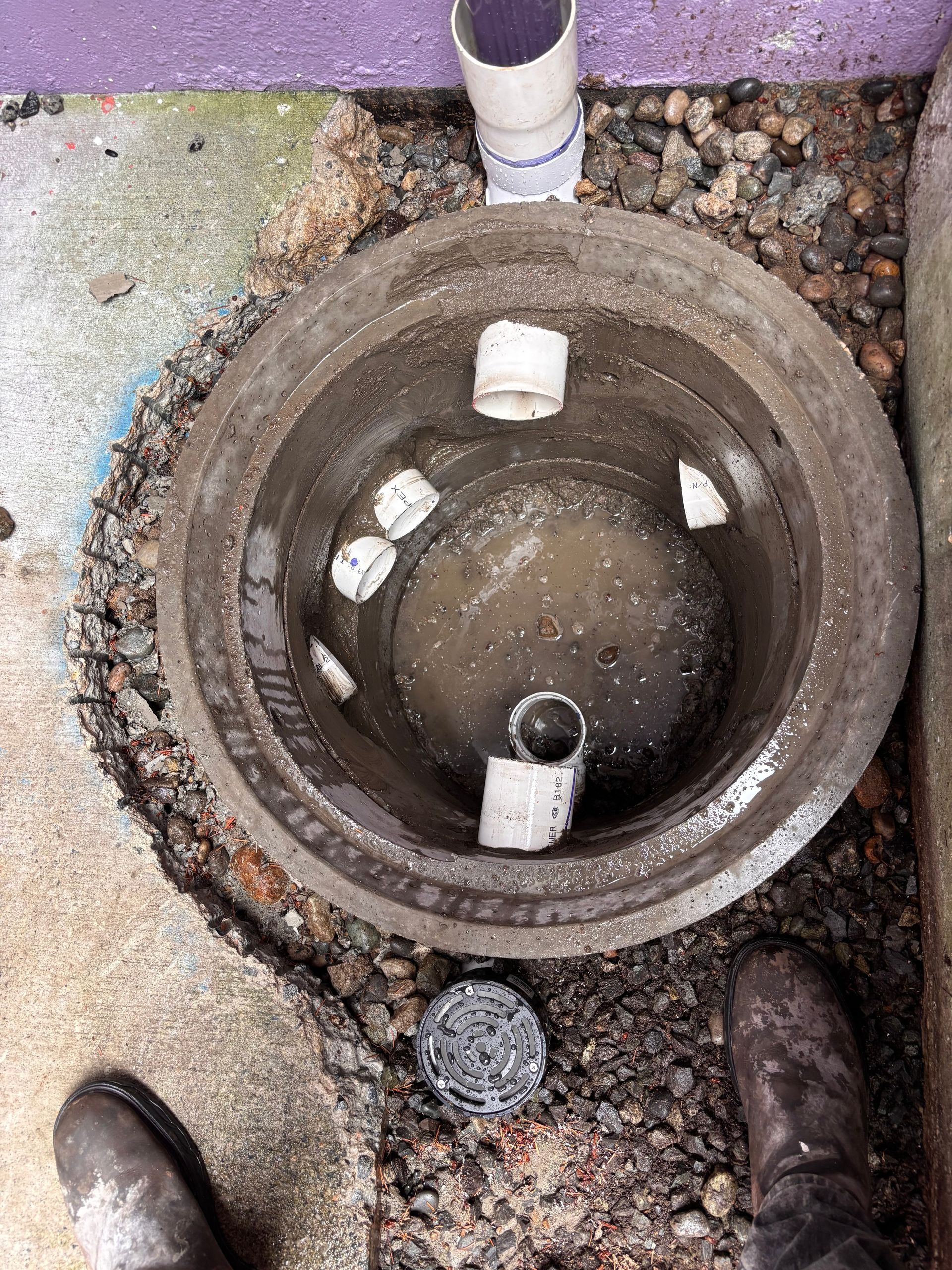 Open replaced sump with water and debris, viewed from above on gravel beside a pipe.