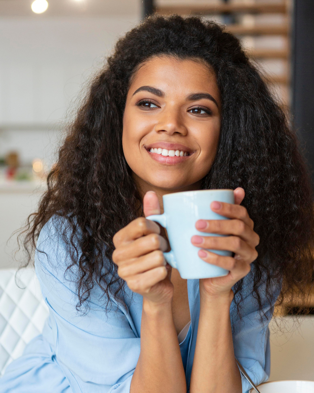 Pessoa sorridente segurando uma caneca azul-clara em uma cozinha ou sala de estar bem iluminada.