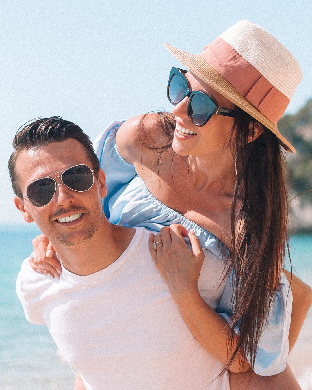 Casal sorridente na praia, a mulher nas costas do homem, ambos usando óculos de sol e chapéus de sol.