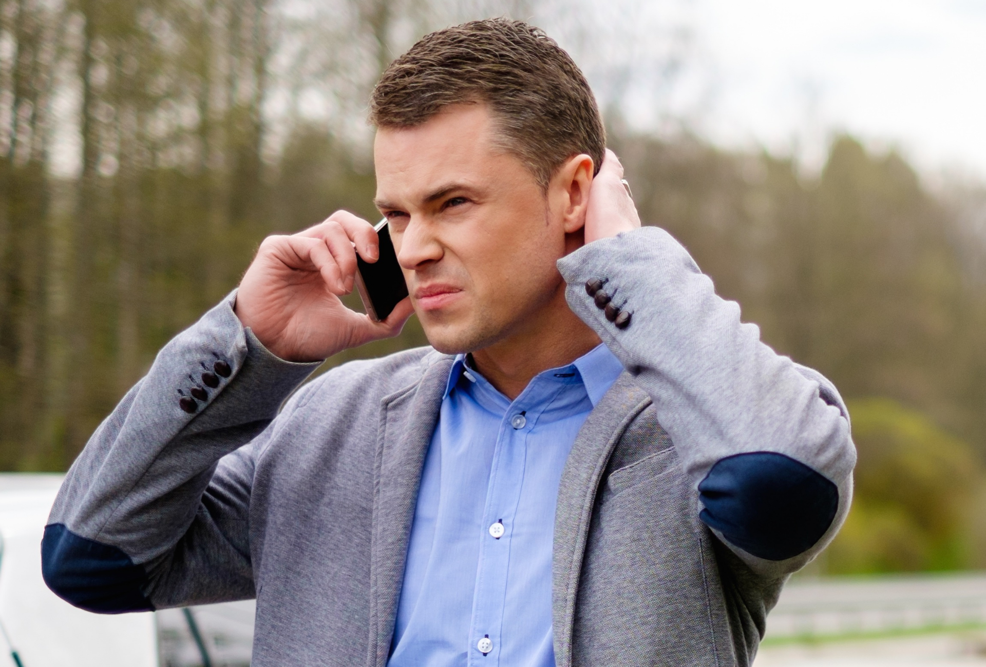 Homem de camisa azul e blazer cinza falando ao telefone ao ar livre, com as duas mãos próximas aos ouvidos.
