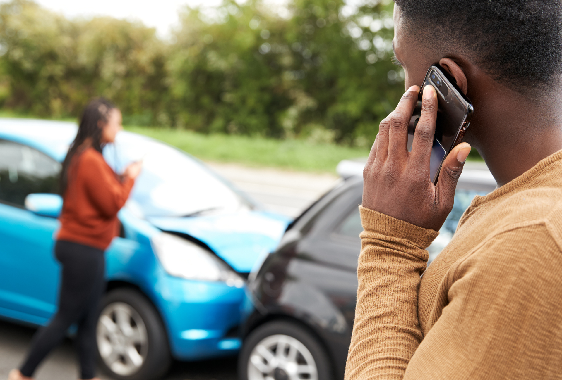 Dois carros envolvidos em uma colisão na beira da estrada, com uma pessoa ao telefone em primeiro plano.
