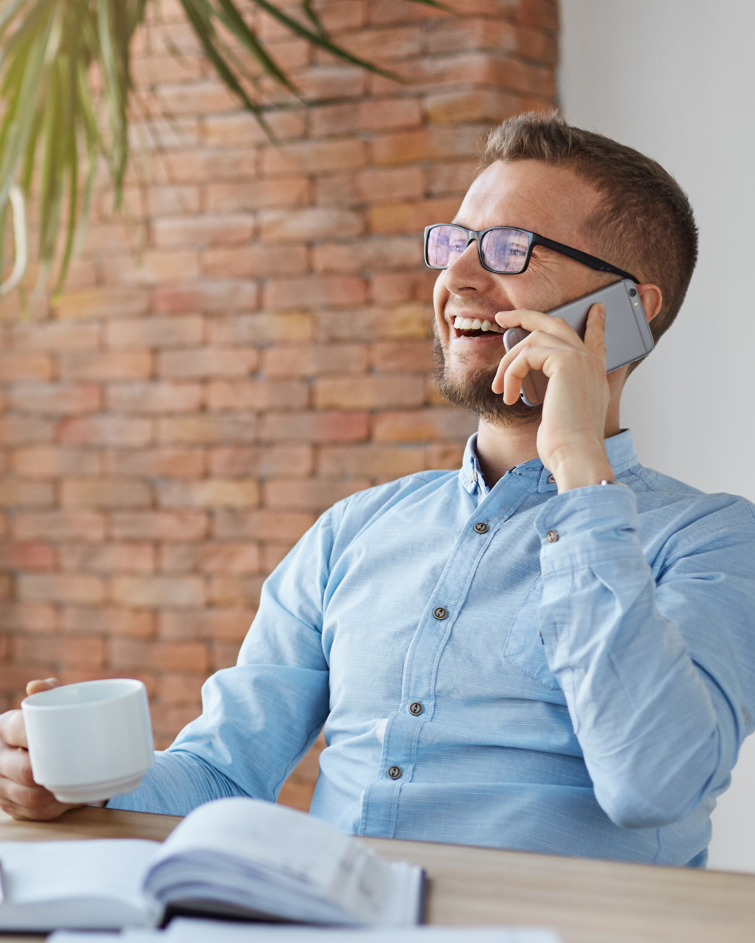 Homem sorridente de camisa azul falando ao telefone enquanto segura uma caneca branca em uma mesa dentro de um ambiente fechado.