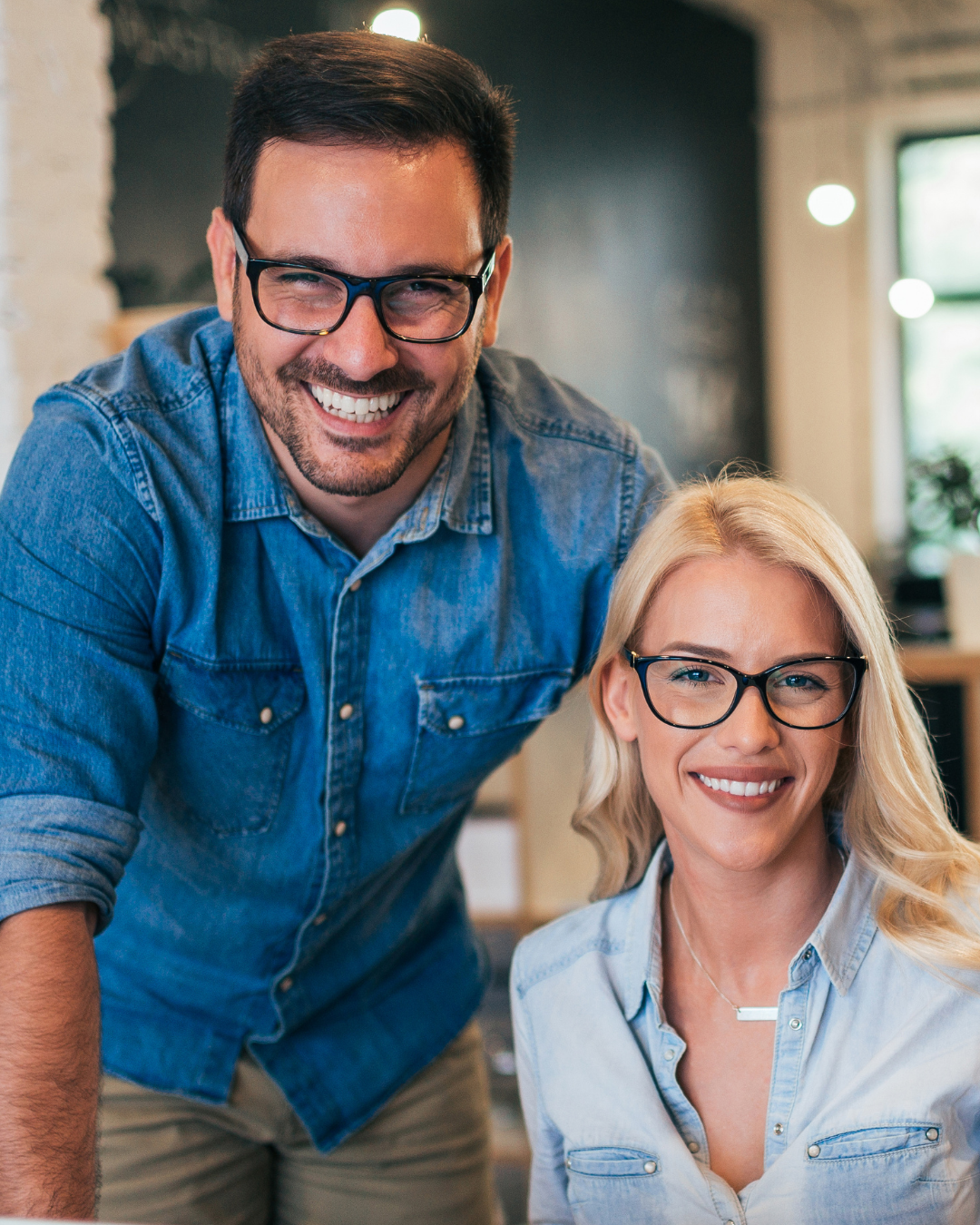 Dois colegas de trabalho sorridentes em um escritório moderno, um atrás do outro, ambos usando óculos.