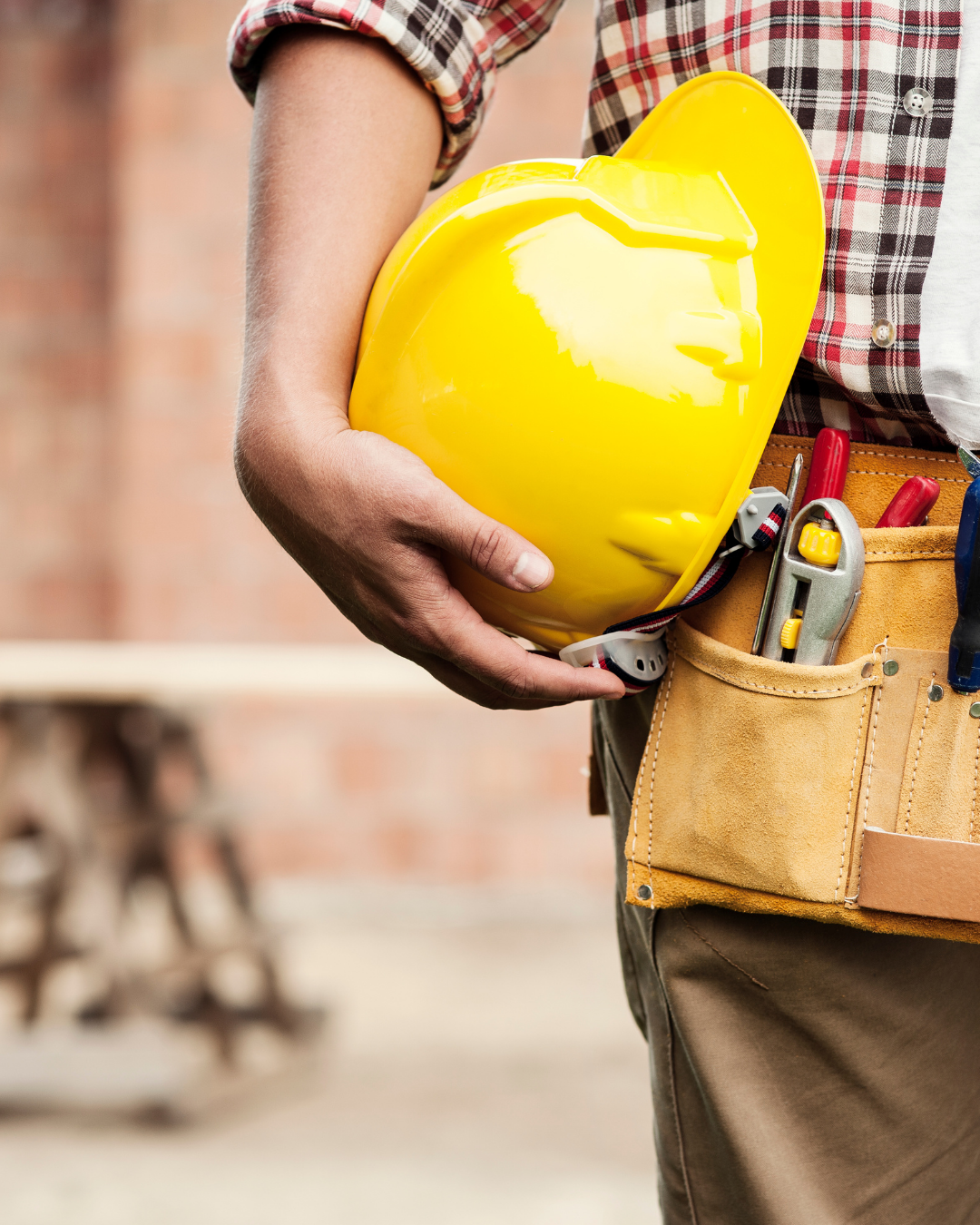 Operário segurando um capacete amarelo ao lado de um cinto de ferramentas em um canteiro de obras.