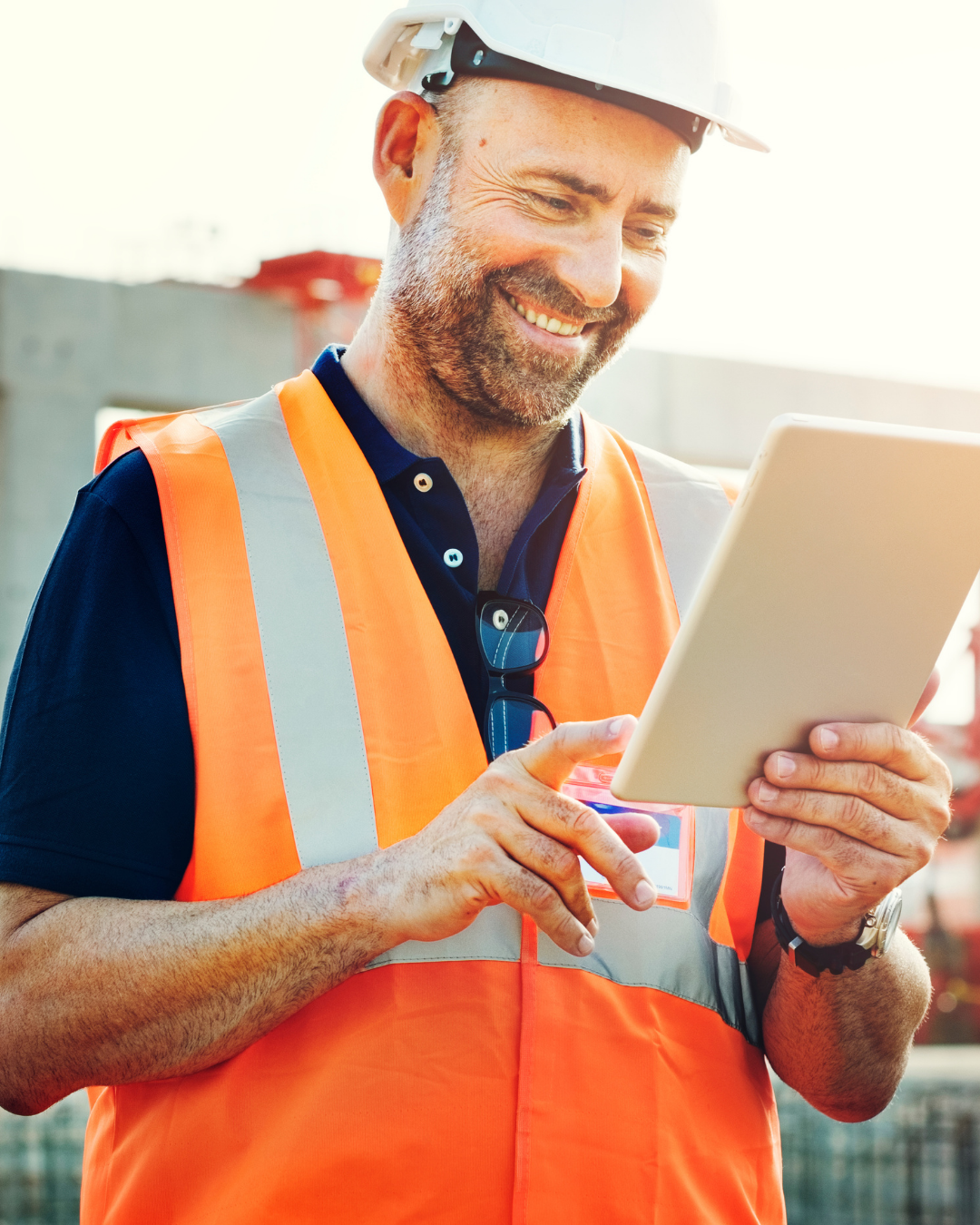 Operário da construção civil com colete de segurança laranja, usando um tablet e sorrindo ao ar livre.