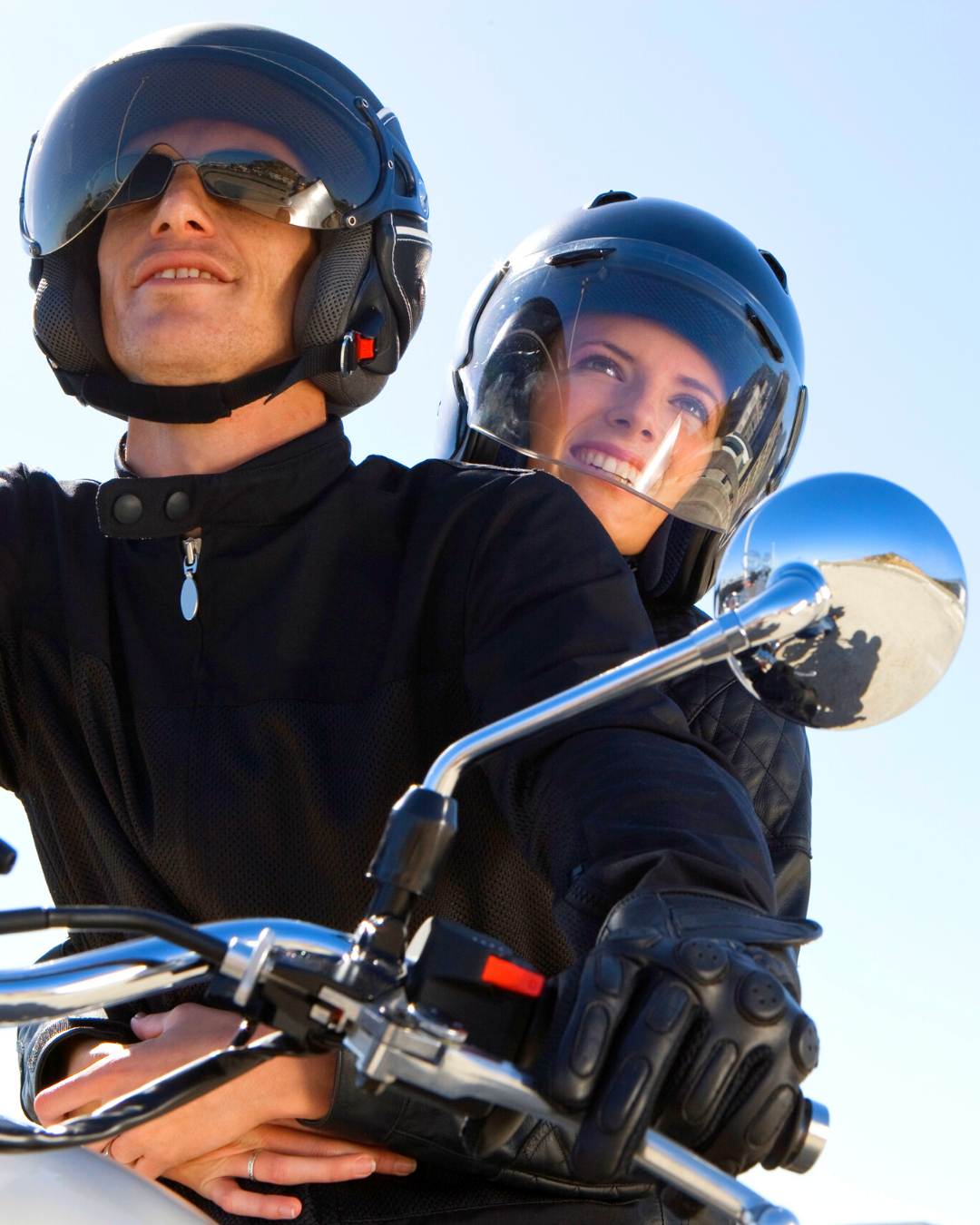 Duas pessoas usando capacetes andando de motocicleta, com um céu azul claro ao fundo.