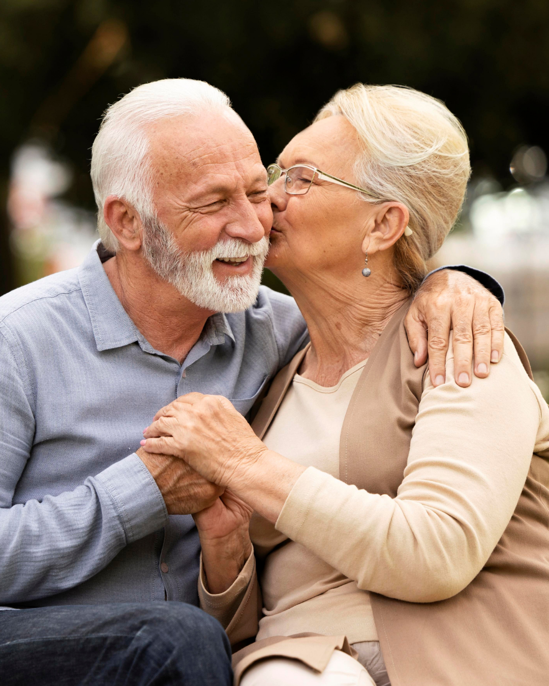 Casal de idosos sorridentes, de mãos dadas e se beijando ao ar livre.