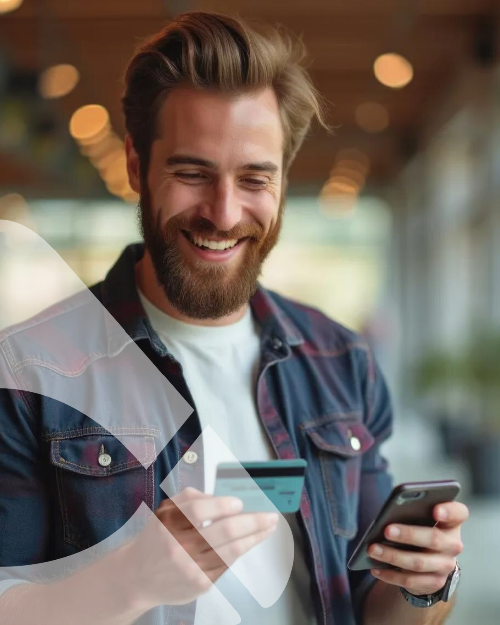 Homem sorridente segurando um cartão de crédito e um smartphone em um café com iluminação suave.