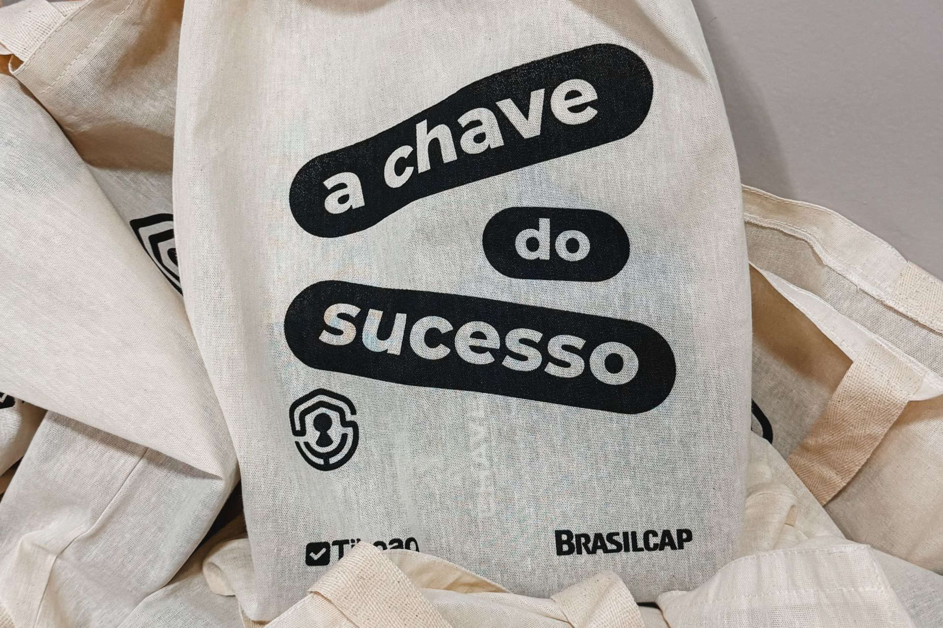 Fabric bags printed with the Portuguese phrase “a chave do sucesso” in black on white fabric.