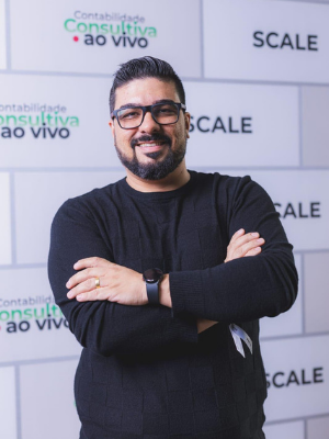 Man in black sweater with glasses, arms crossed, standing before a branded SCALE backdrop
