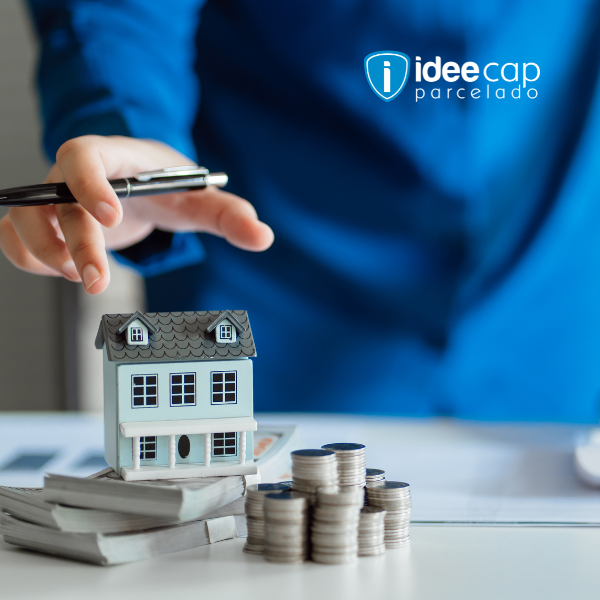 Mão segurando uma caneta acima de uma casa em miniatura, moedas empilhadas e documentos sobre uma mesa, com o logotipo da Ideecap.