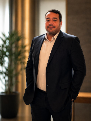 Man in a dark suit standing indoors beside a potted plant and curtains