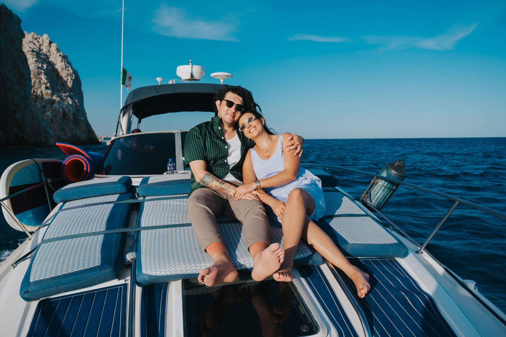 Casal sentado no convés de um iate com o oceano e os penhascos rochosos ao fundo.