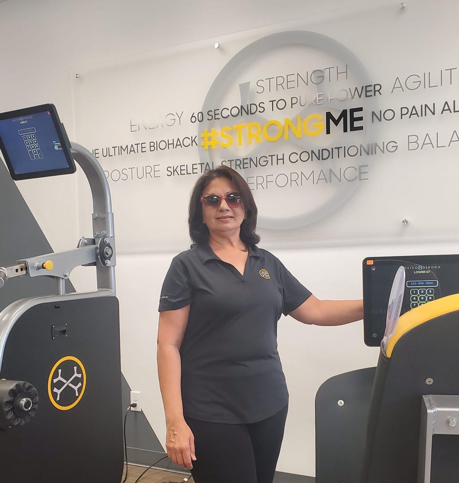 Woman in sunglasses standing in a gym beside exercise equipment and a wall with strength-training words