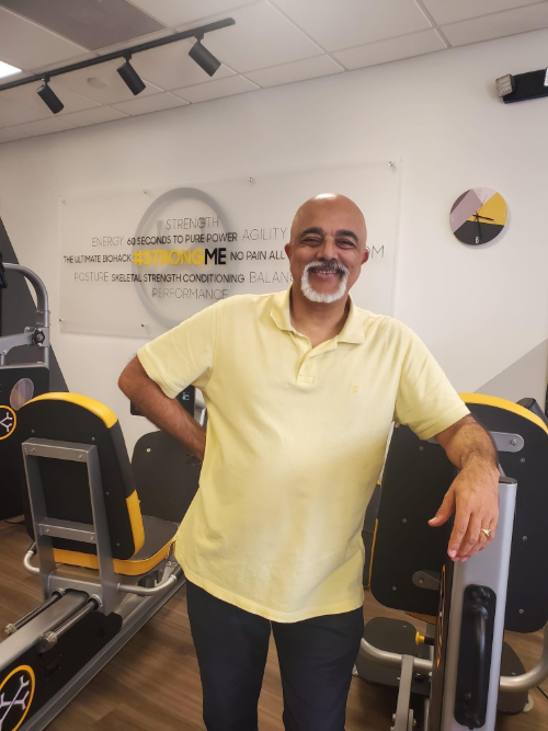 Man standing in a gym beside workout machines, wearing a pale yellow polo shirt and smiling.