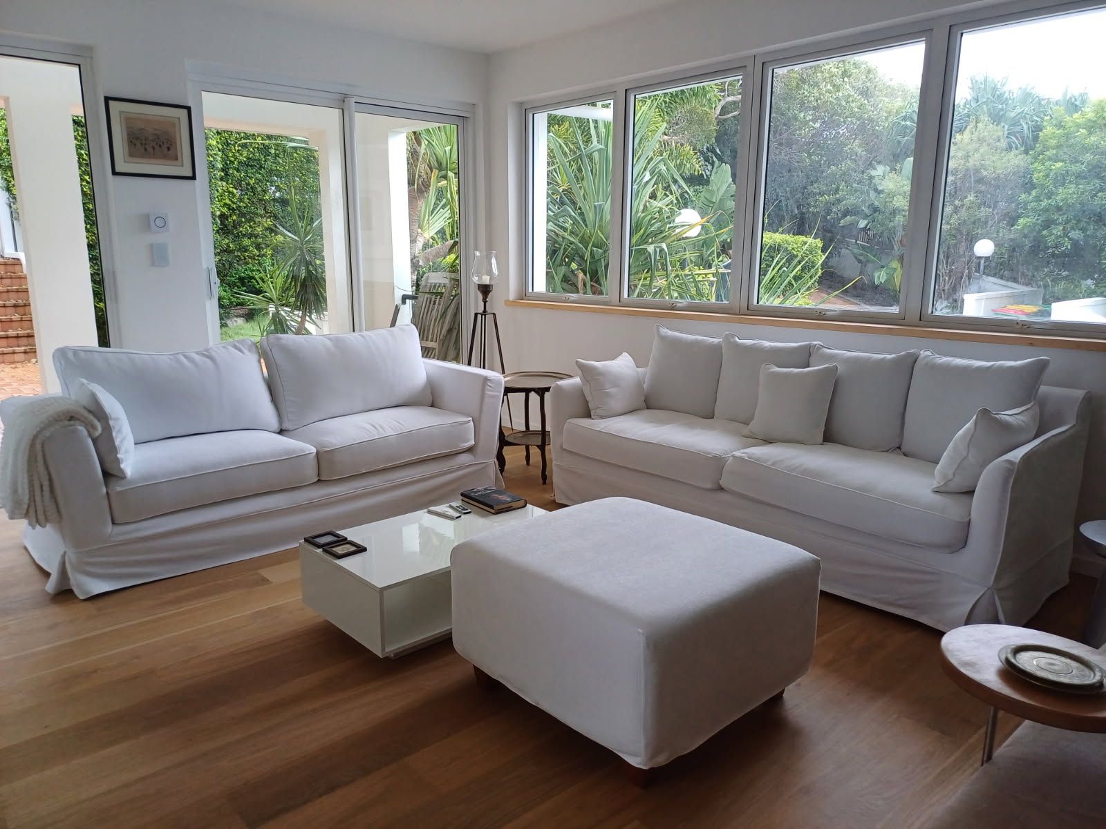 A bright living room features white sofas and an ottoman on hardwood floors, with large windows overlooking greenery.