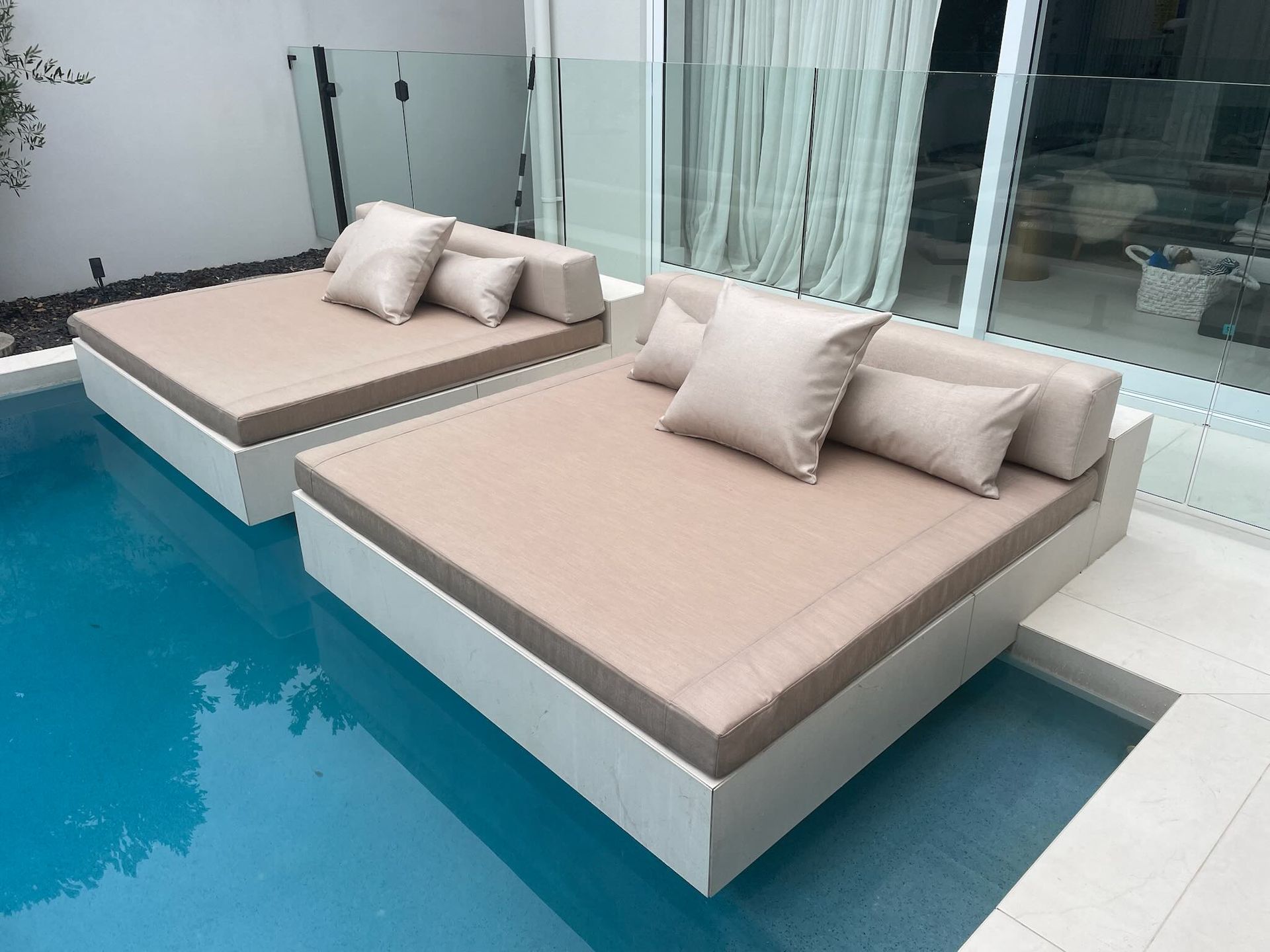 Two beige, square daybeds floating on a swimming pool next to a modern building with a glass fence.