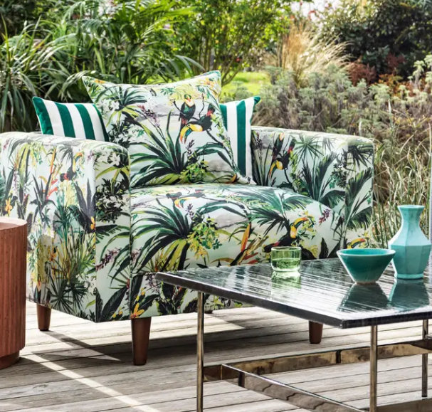 A patterned armchair with tropical plant and bird motifs, featuring striped pillows on a deck beside a glass patio table.