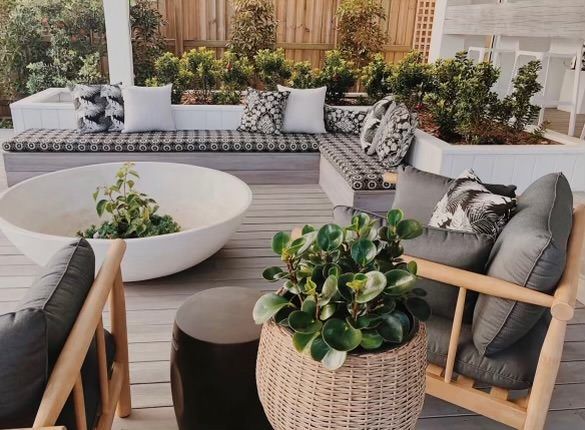 Modern outdoor patio lounge with white sectional seating, wooden chairs, a large circular planter, and lush greenery.