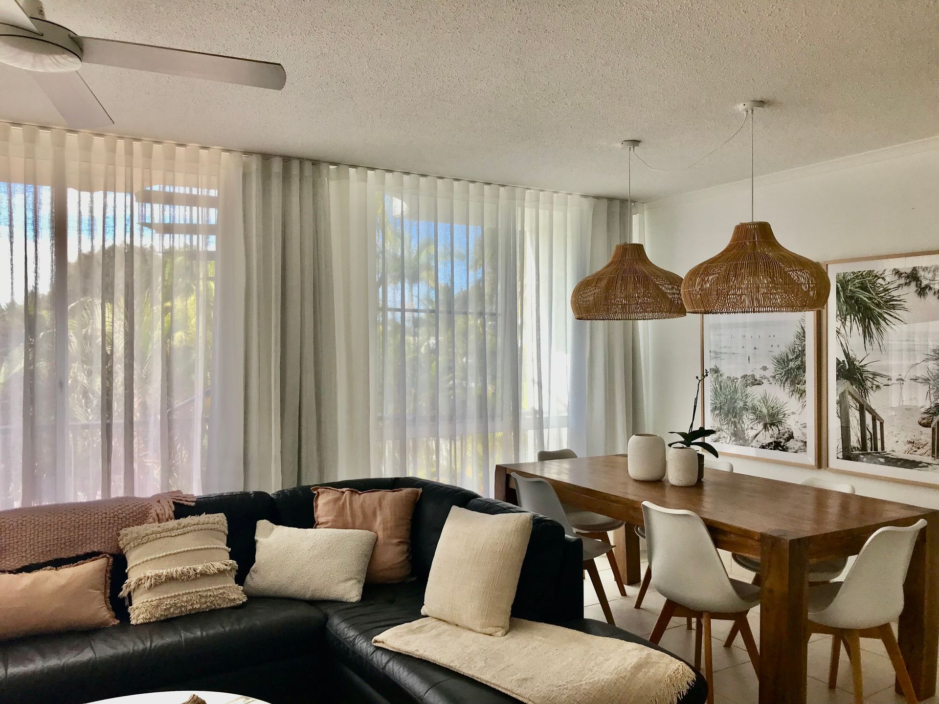 A bright living and dining area with a dark sectional sofa, a wooden dining table, white chairs, and sheer curtains.