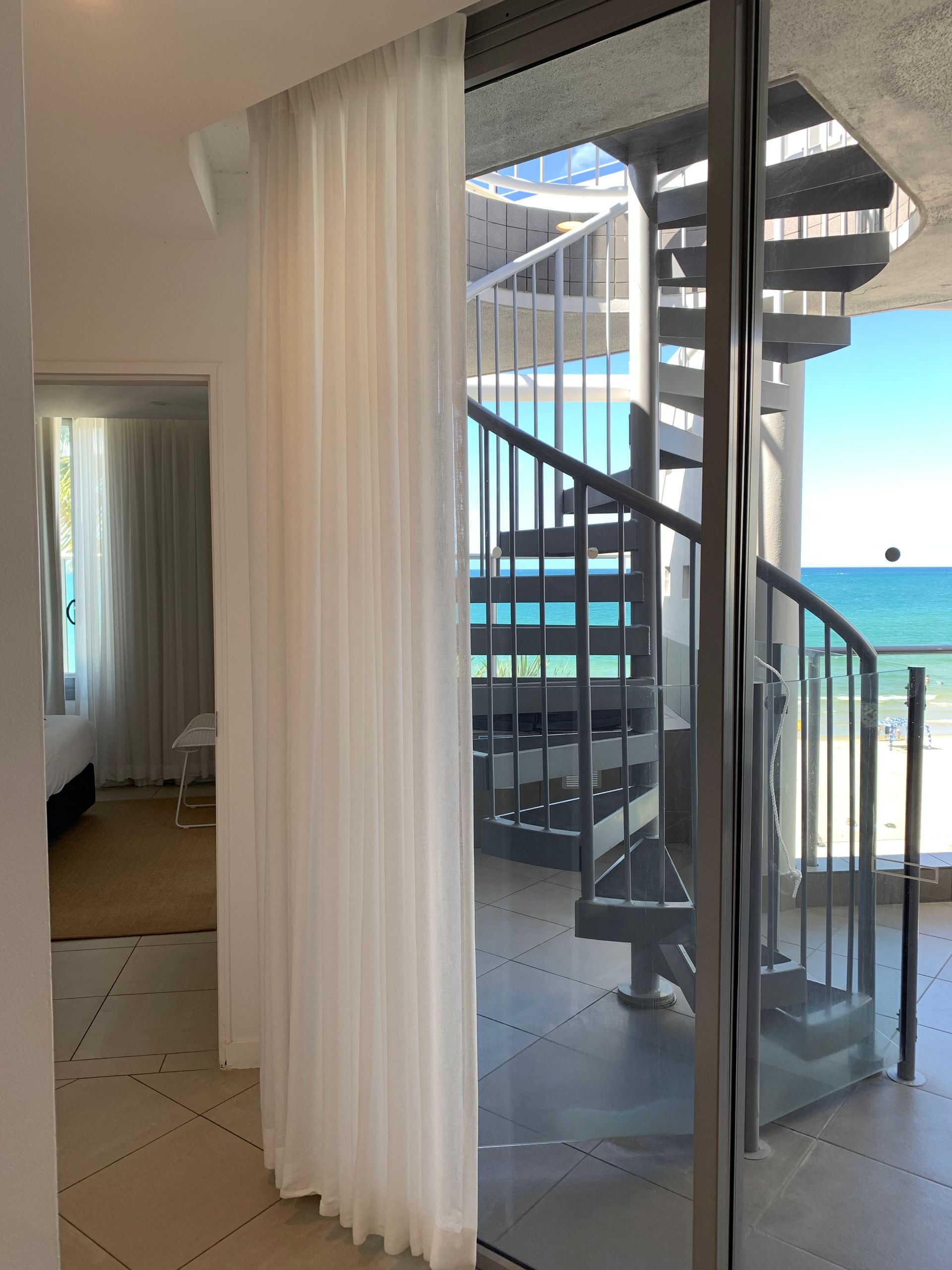 A spiral staircase outside a glass balcony door overlooks a sunny beach, with a sheer curtain inside the room.