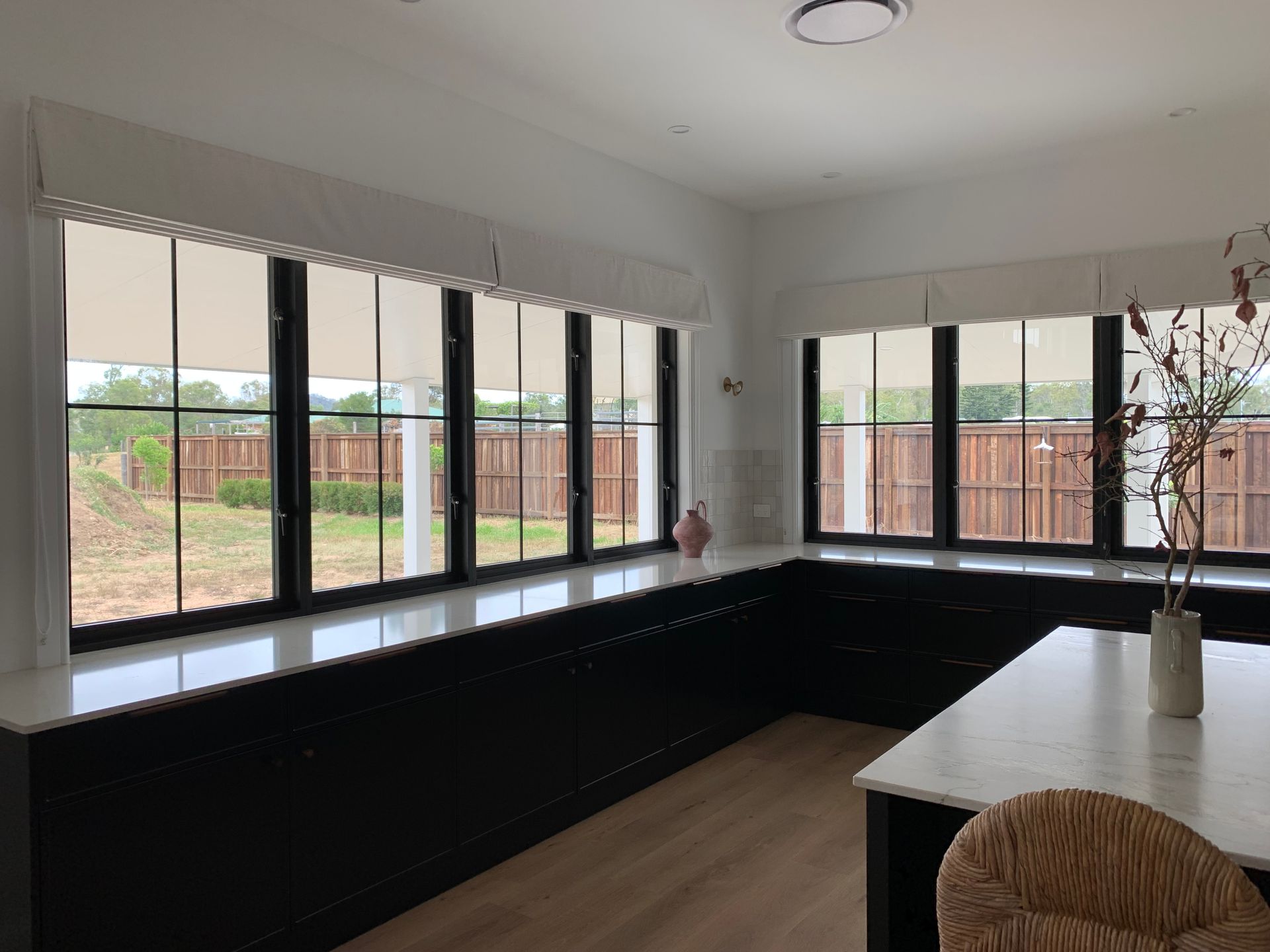 Modern kitchen with black cabinetry, long white countertops under large windows with cream shades, and a wood-look floor.