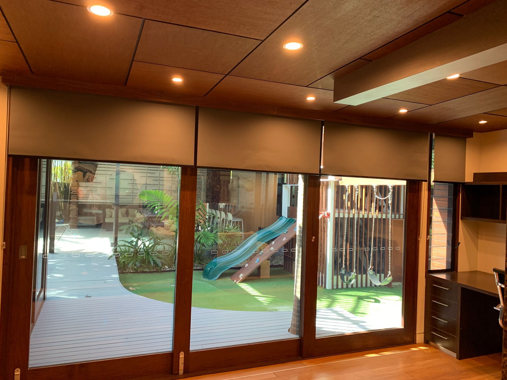 View from an indoor room through large glass sliding doors to a backyard with a playground, slide, and artificial lawn.