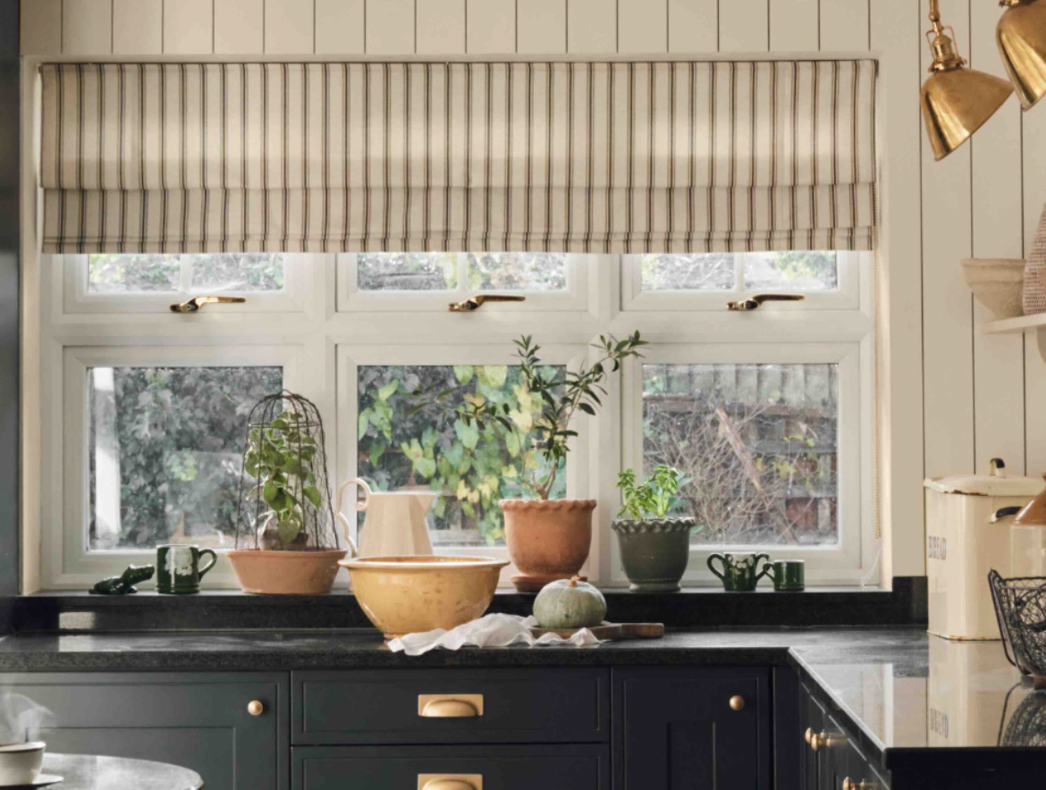 A cozy kitchen window with a striped roman shade, potted plants, and a mixing bowl on a dark countertop.