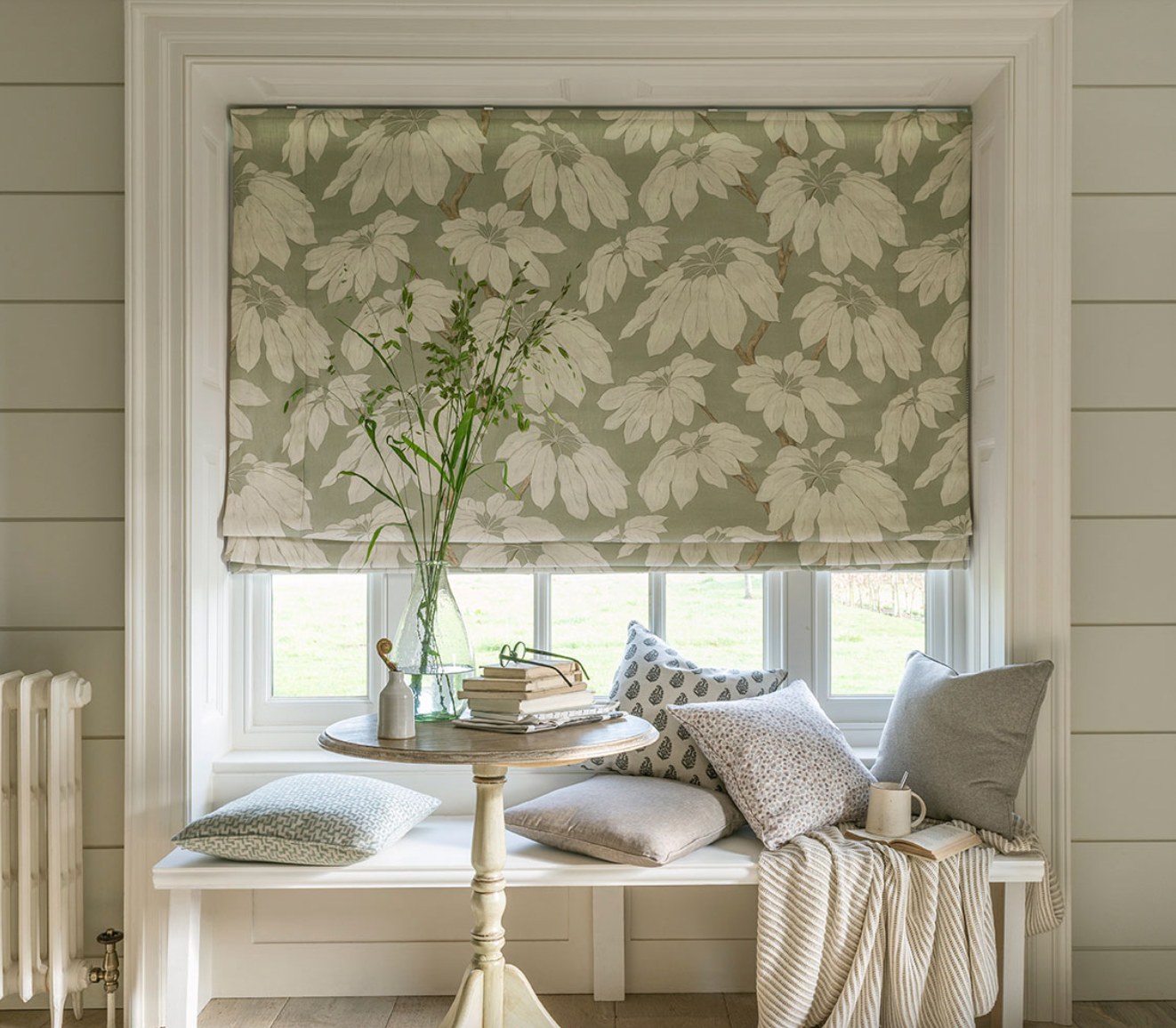 A cozy window seat with floral Roman shades, a small table, several cushions, and a throw blanket in a rustic room.