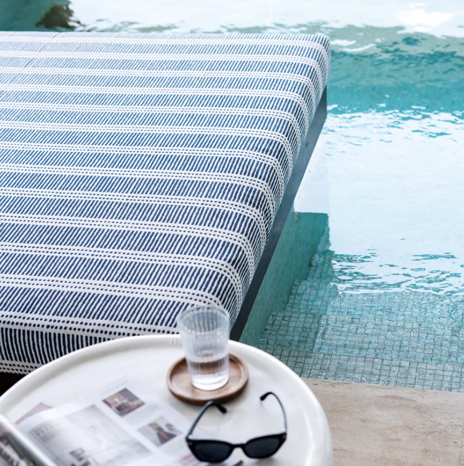 A white table with a glass of water, sunglasses, and a magazine next to a poolside lounge chair with a striped cushion.