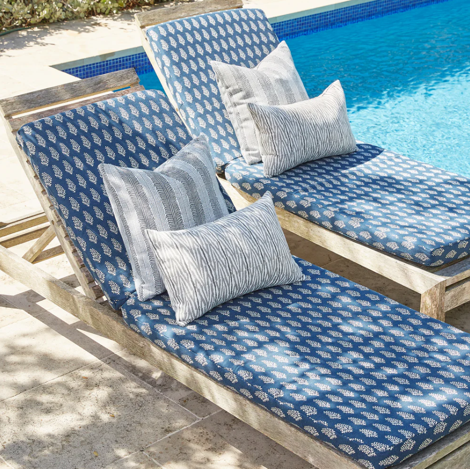 Two wooden lounge chairs with blue patterned cushions and patterned throw pillows beside a sunlit swimming pool.