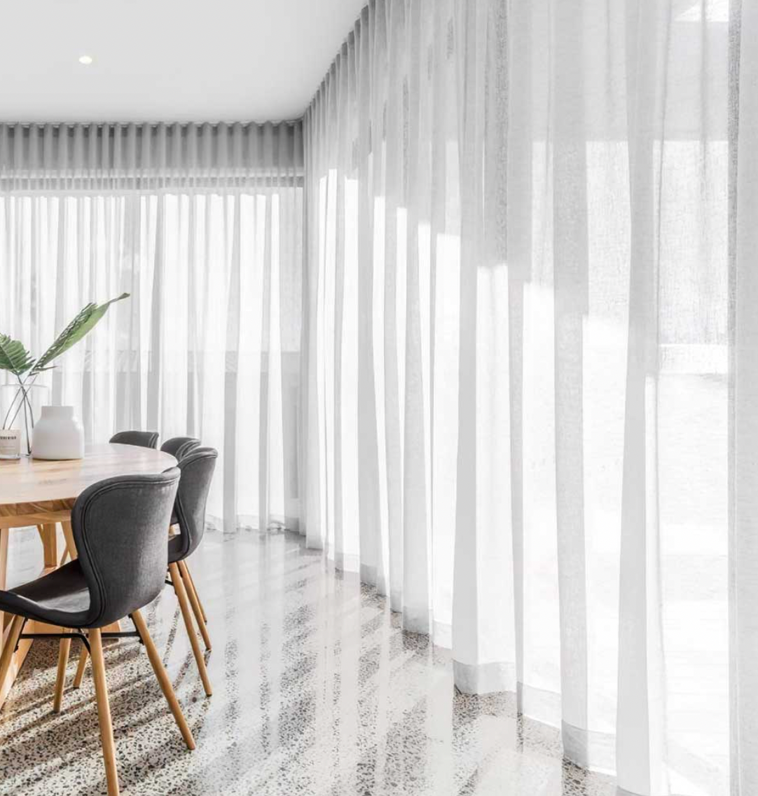 A dining room with dark gray chairs, a wooden table, and floor-to-ceiling white sheer curtains over large glass windows.