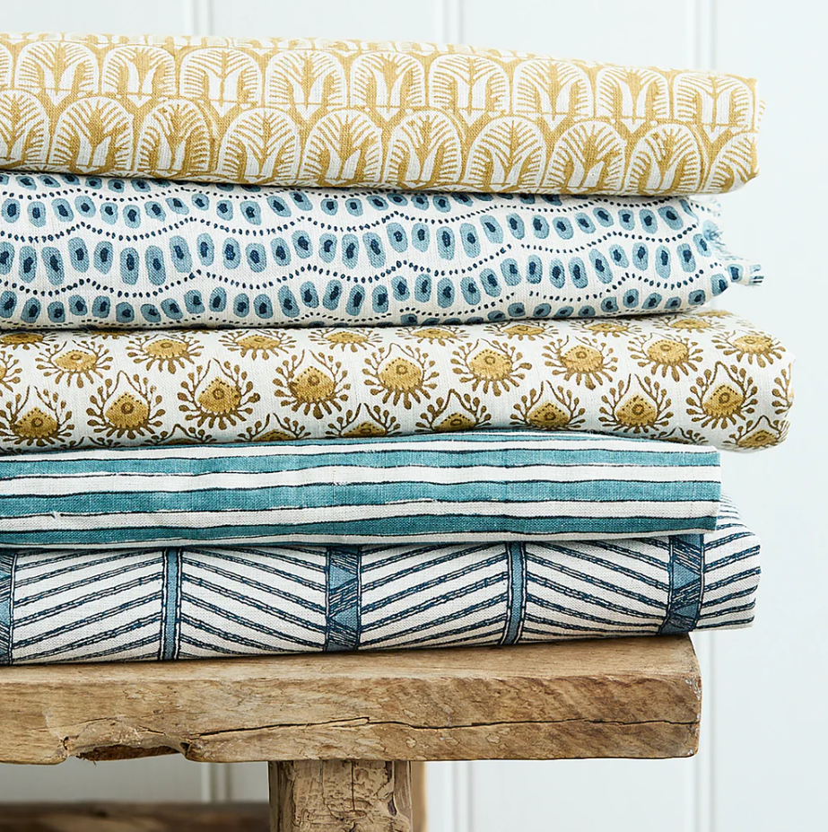 A stack of five folded fabrics with various patterns and colors—gold, blue, and white—sitting on a rustic wooden bench.