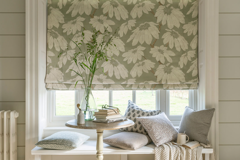 A sunlit window seat featuring floral roman shades, stacked pillows, and a glass vase on a wooden side table.