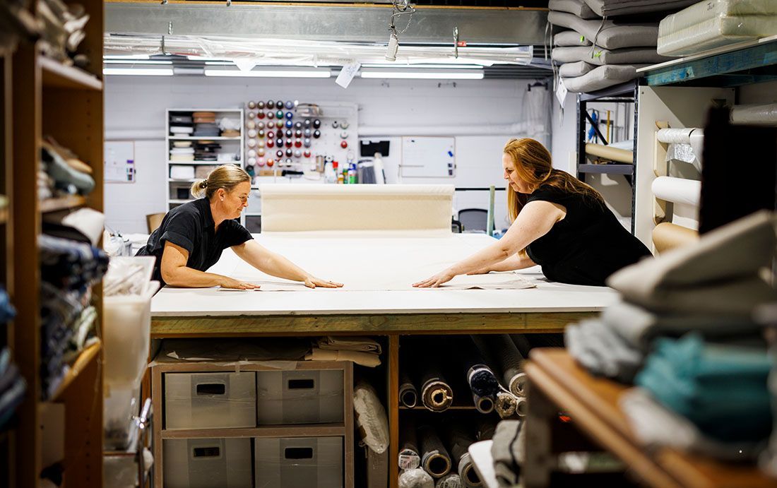 Two people lean over a large cutting table in a textile workshop, smoothing out a long length of white fabric.