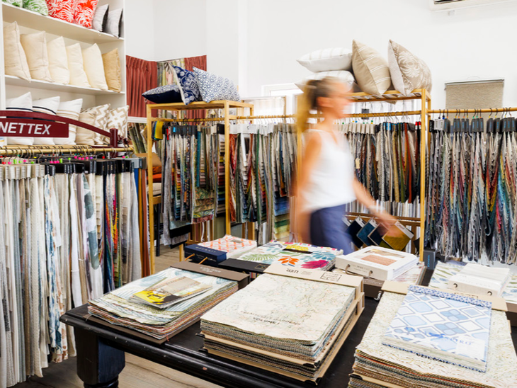 A person walking through a fabric store filled with shelves of textile samples, throw pillows, and stacks of swatch books.
