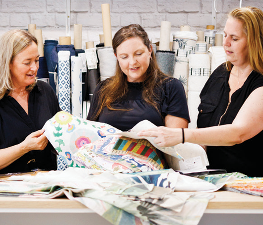Three people discuss various fabric swatches at a table in a studio filled with rolls of textiles.
