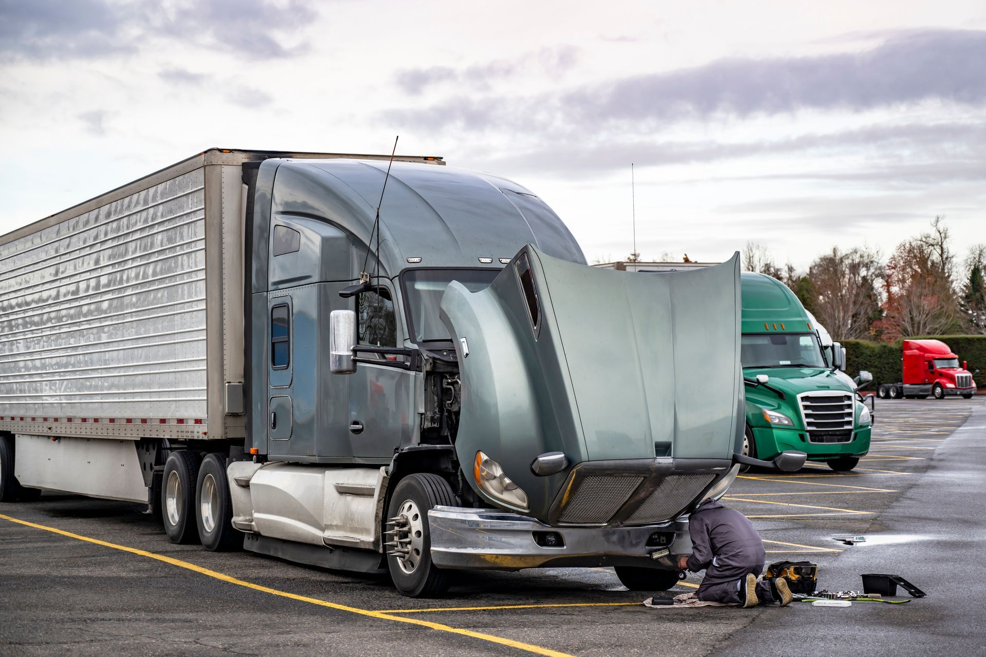 A truck driver repairing a broken big-rig semi-truck with an open hood outdoors.