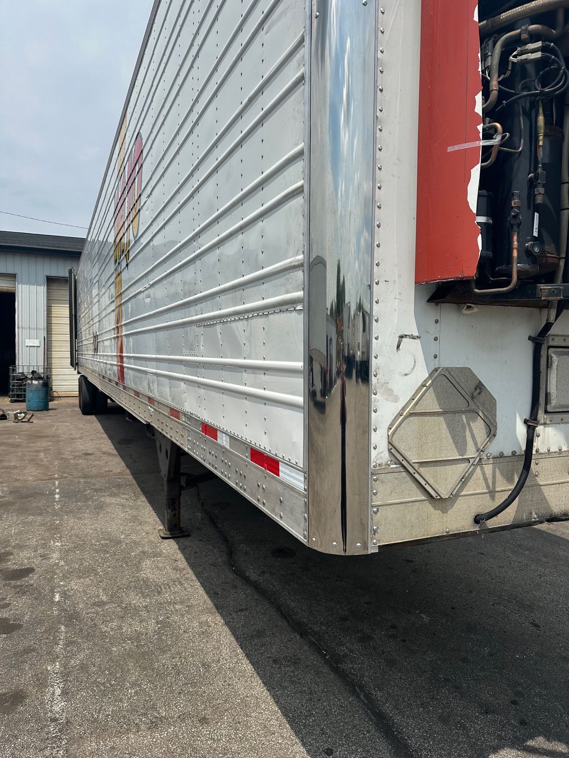 A side view of a parked semi-trailer with a metallic finish, red accents, and a refrigeration unit at the front.