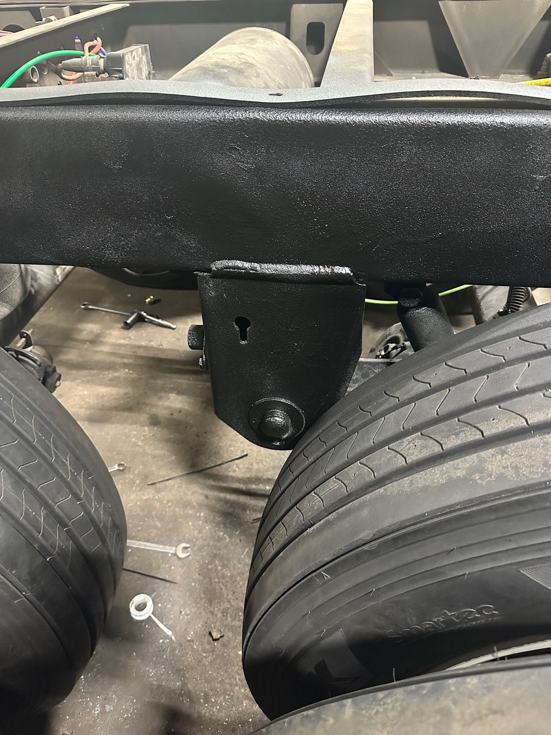 A close-up of a black metal suspension hanger welded to a truck frame between two large vehicle tires.