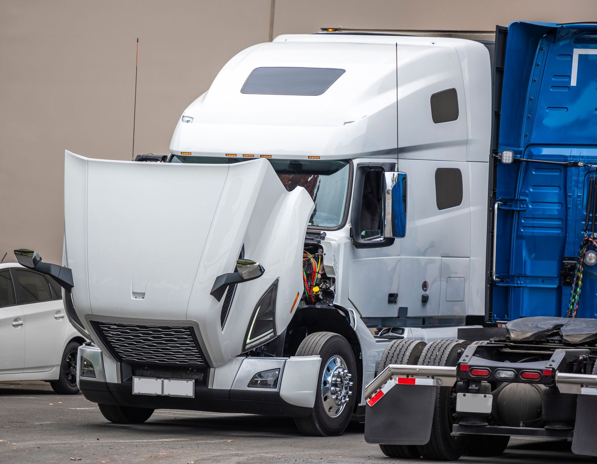 White semi truck with raised hood undergoing engine repair in parking lot beside blue trailer depot.