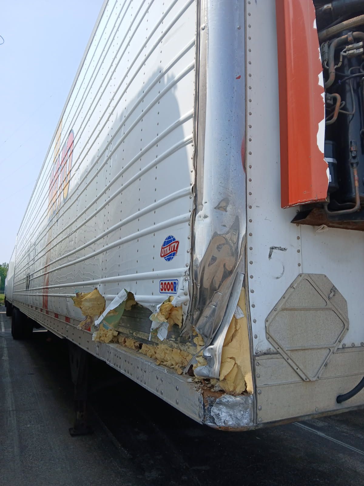 Close-up of a damaged, dented metal side panel on a refrigerated semi-trailer, revealing exposed yellow insulation foam.