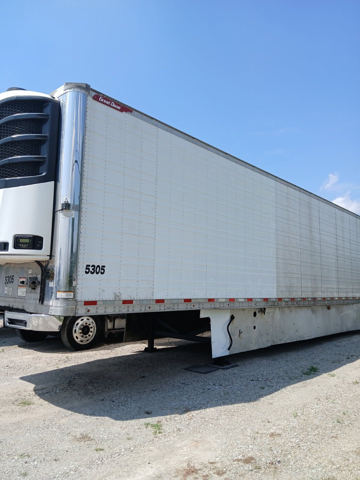 A white refrigerated semi-trailer parked on a gravel lot under a clear blue sky.