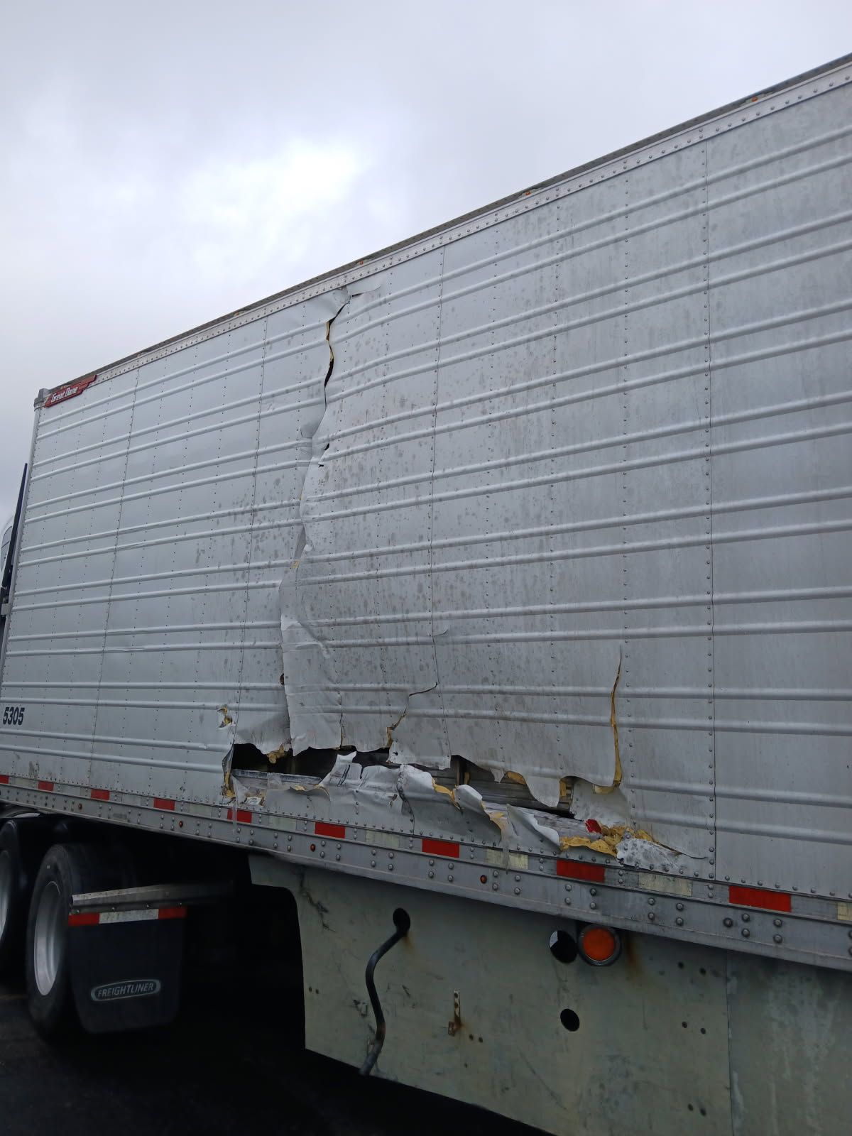 A damaged silver semi-trailer with a large vertical tear and buckled metal paneling on its side.