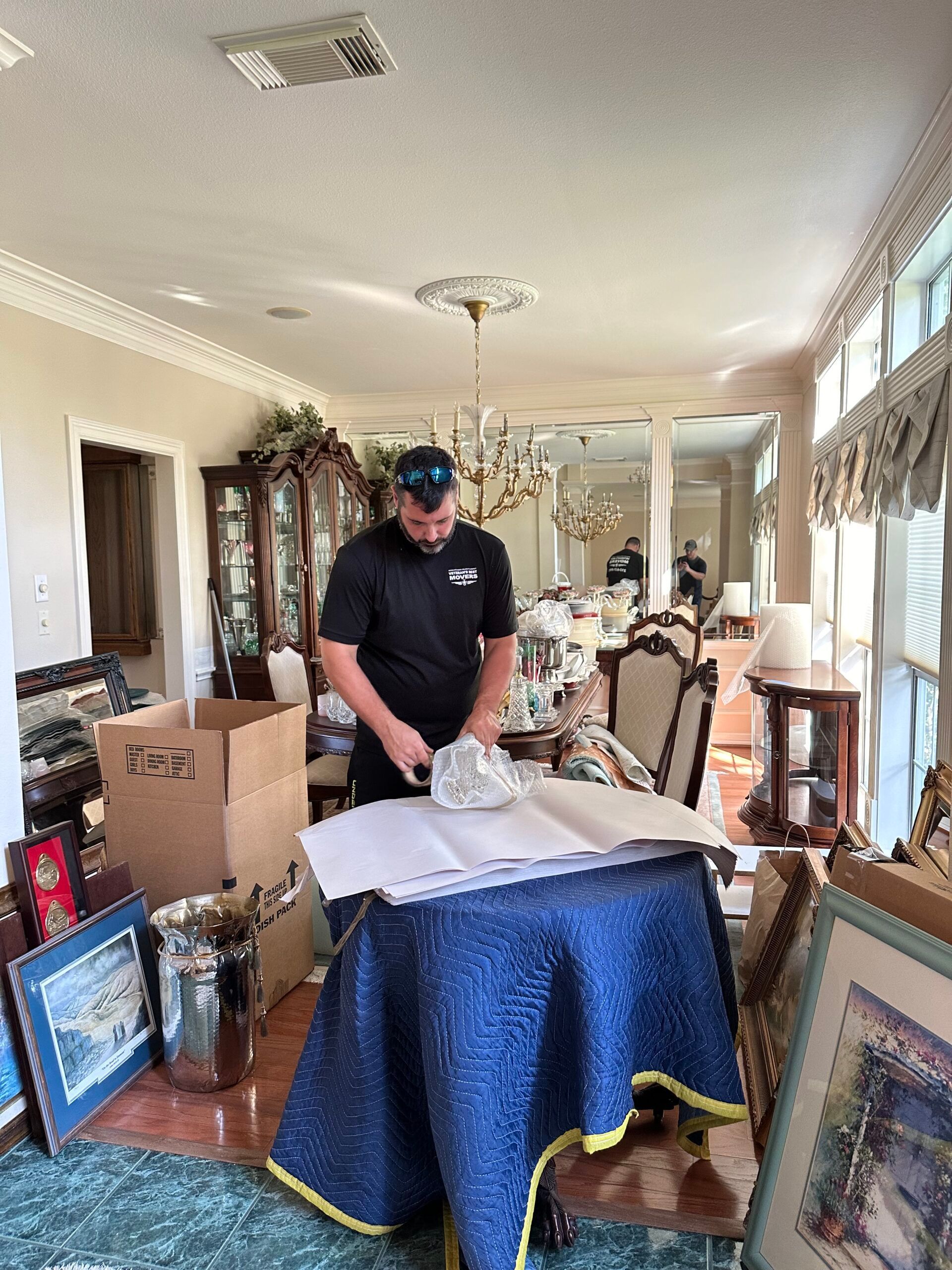 Man wrapping object on a table, preparing for move, surrounded by furniture and boxes.